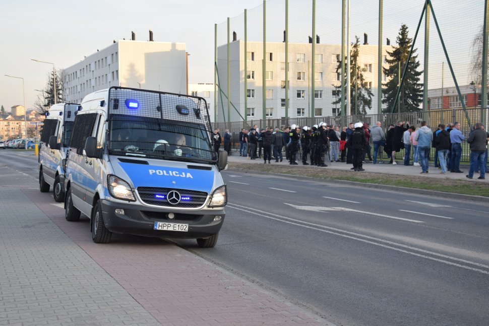 zdjęcie przedstawia policjantów i kibiców stojących przed stadionem piłkarskim. W tle widać bloki mieszkalne. Z lewej strony zdjęcia widać policyjne radiowozy