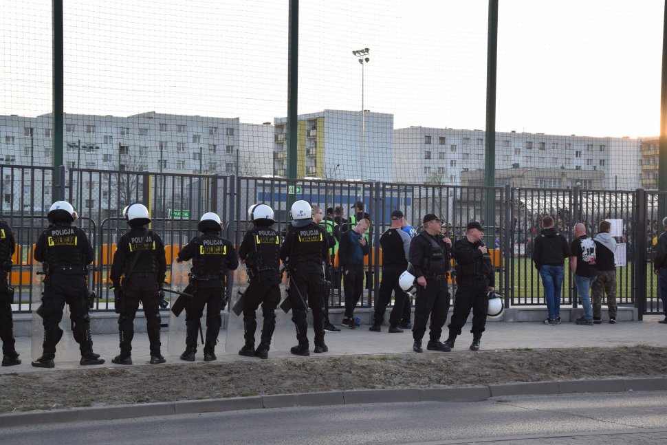 zdjęcie przedstawia policjantów i kibiców stojących przed stadionem piłkarskim. W tle widać bloki mieszkalne