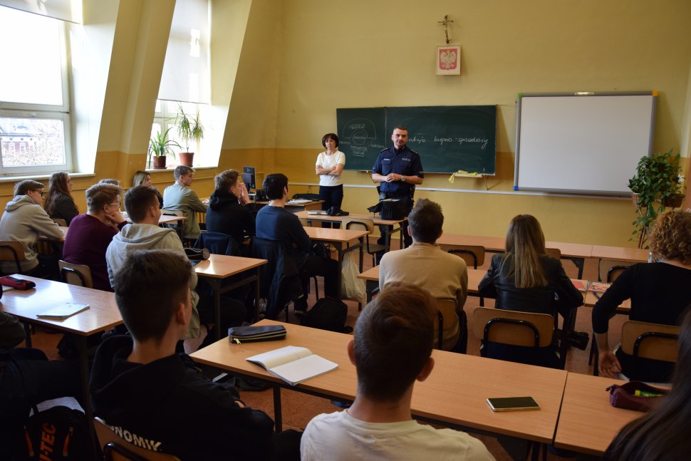 zdjęcie przedstawia szkolną klasę, w której uczniowie siedzą w ławkach. Rozmawia z nimi policjant 