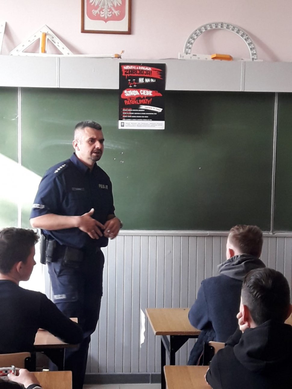 na zdjęciu widać szkolną klasę, w której policjant stojący przy tablicy, rozmawia z uczniami siedzącymi w ławkach
