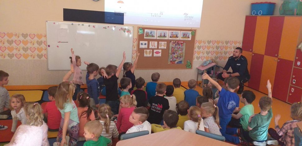 na zdjęciu widać szkolną klasę, w której policjant rozmawia z uczniami siedzącymi na podłodze 