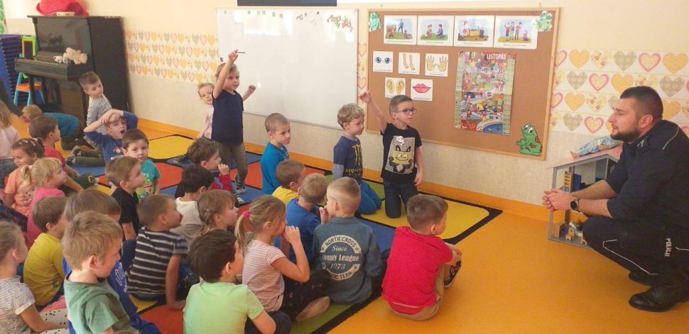 na zdjęciu widać szkolną klasę, w której policjant rozmawia z uczniami siedzącymi na podłodze 