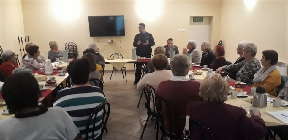 na zdjęciu widać salę, na której przy stołach siedzą seniorzy i rozmawia z nimi policjant  