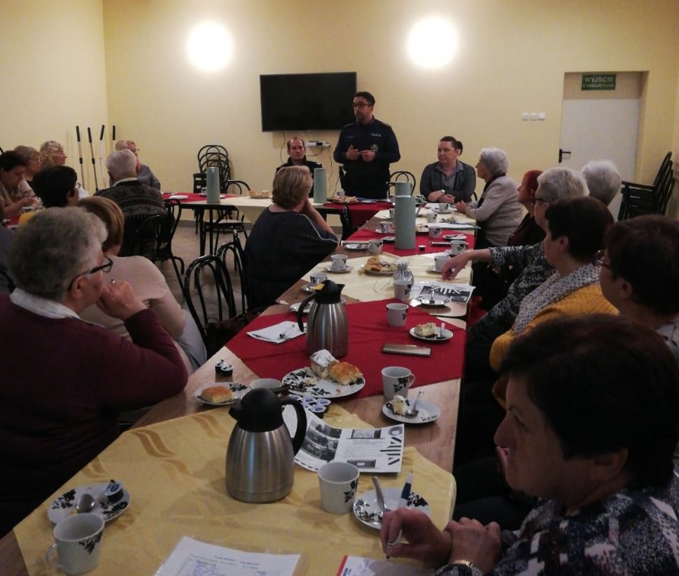 na zdjęciu widać salę, na której przy stołach siedzą seniorzy i rozmawia z nimi policjant  