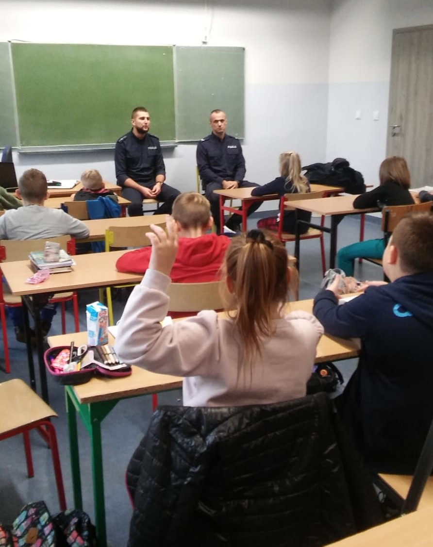 na zdjęciu widać szkolną klasę, w której dzielnicowi siedzący przy tablicy, rozmawiają z uczniami siedzącymi w ławkach