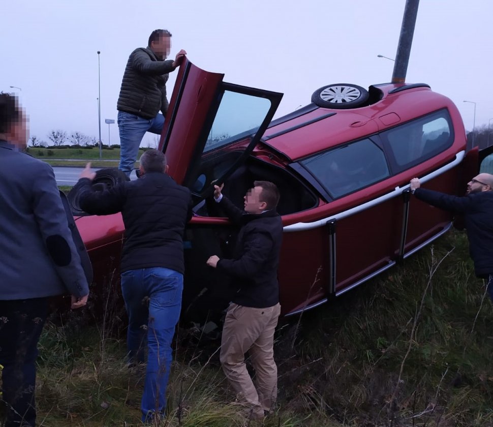 Na fotografii widać leżący na boku w rowie samochód osobowy. Na samochodzie stoi mężczyzna który trzyma otwarte drzwi  od strony pasażera. Kolejni mężczyźni trzymają samochód, aby nie przewrócił się na dach. 