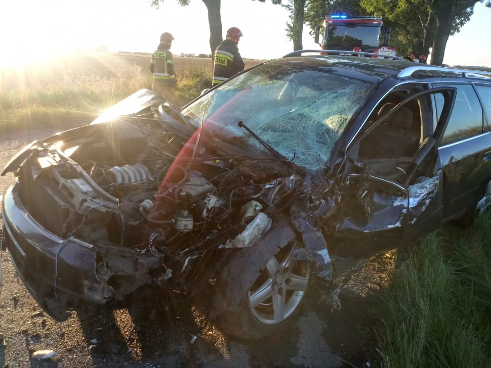 Na fotografii widać zniszczony samochód osobowy. W tle zdjęcia widać strażaków 