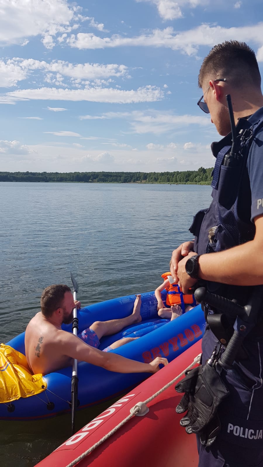 Na zdjęciu widać zalew, po którym na pontonie pływa policjant i obserwuje mężczyznę i dziecko pływających na innym pontonie . Na innym pontonie   