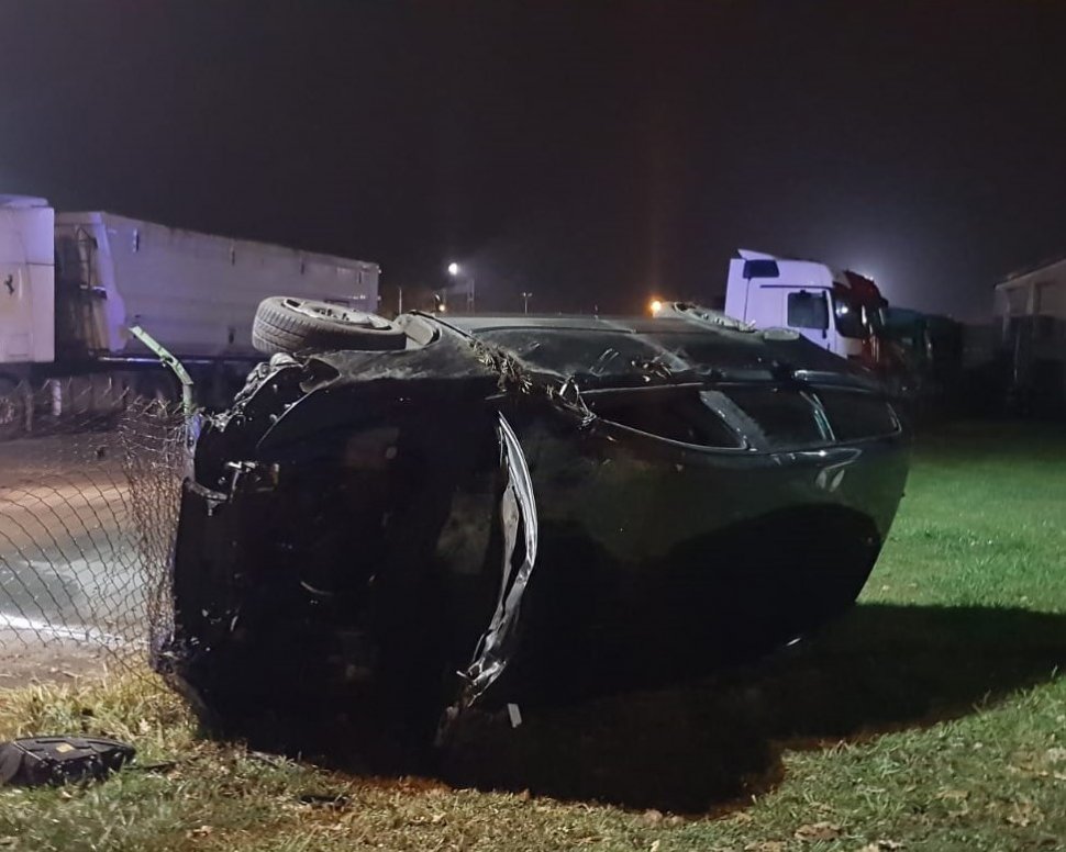 Na zdjęciu widać uszkodzony samochód osobowy, który uderzył w ogrodzenie i leży na boku. W tle zdjęcia widać zaparkowane samochody ciężarowe.  