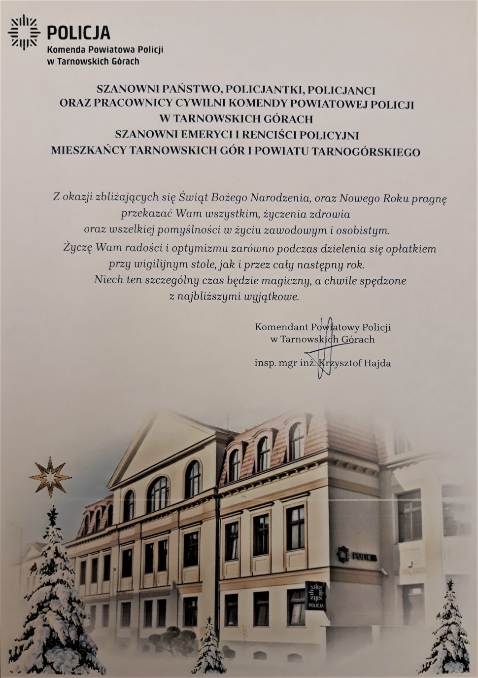 Zdjęcie przedstawia grafikę: budynek Komendy Policji w Tarnowskich Górach, przy którym stoją ośnieżone choinki. Nad grafiką widnieje tekst: życzenia Komendanta Powiatowego Policji w Tarnowskich Górach 
