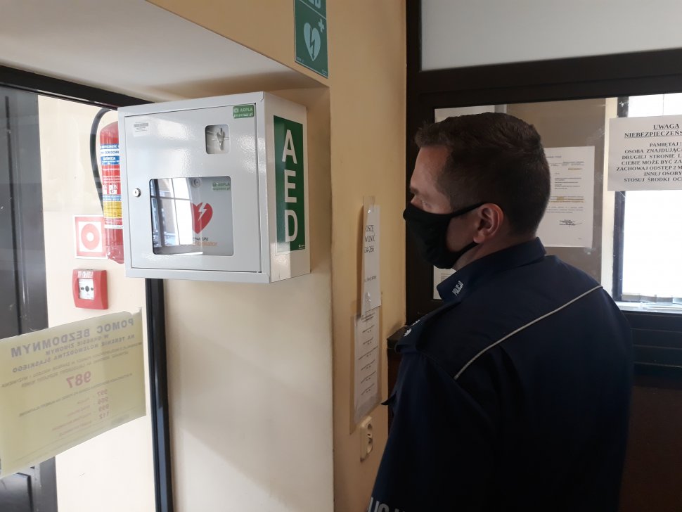 Zdjęcie przedstawia policjanta stojącego obok Automatycznego Defibrylatora Zewnętrznego