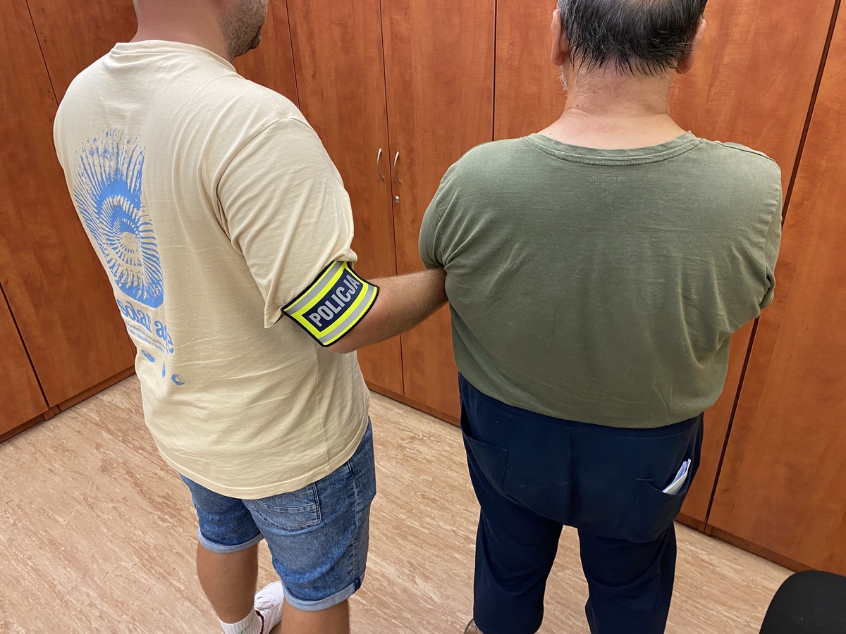 policjant z opaską trzyma za rękę zatrzymanego w pomieszczeniu