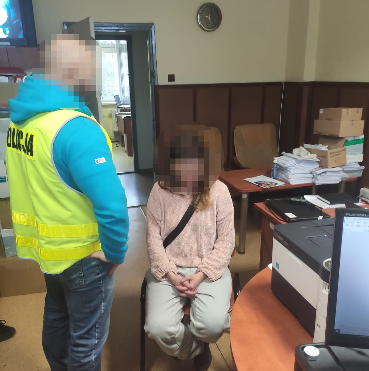 na zdjęciu policjant w odblaskowej kamizelce, stoi przy zatrzymanej kobiecie, która siedzi przy biurku
