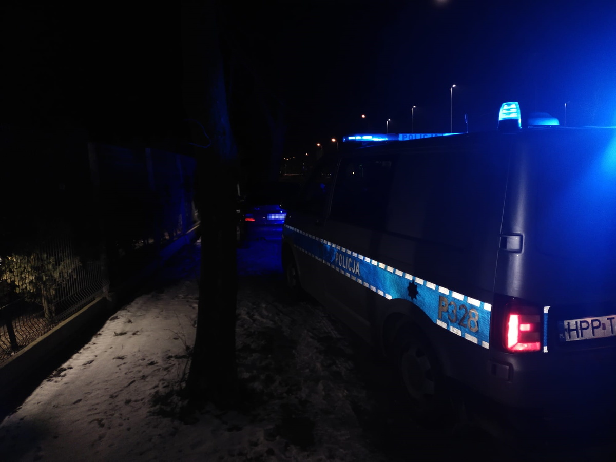 na zdjęciu policyjny radiowóz z nocnej aurze, z włączonymi sygnałami błyskowymi, na drodze, podczas stojącego przed nim, kontroli pojazdu