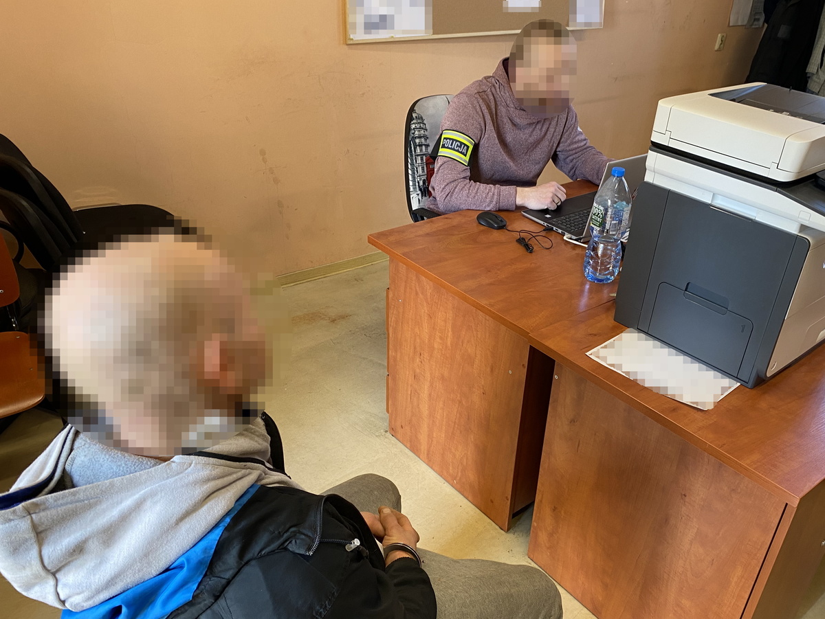 na zdjęciu zatrzymany siedzi przy biurku, za którym siedzi policjant w ubiorze cywilnym z opaska na ramieniu z napisem &#039;policja&#039;
