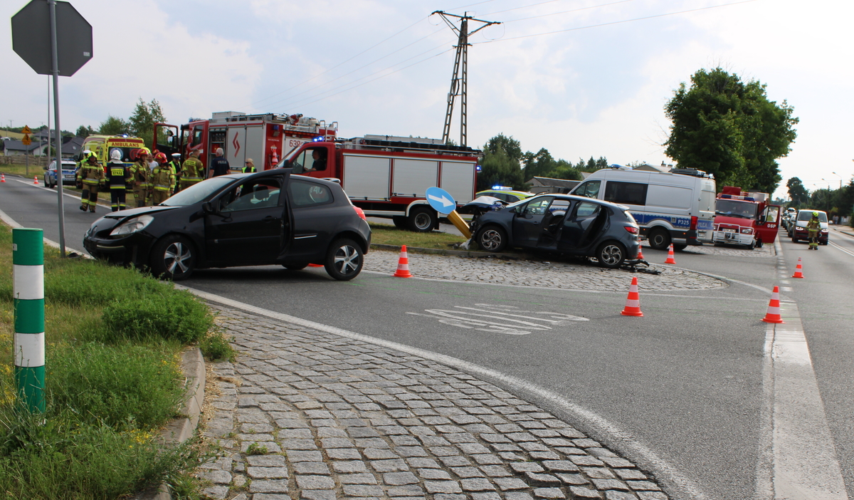 na zdjęciu dwa samochody osobowe na skrzyżowaniu, z widocznymi uszkodzeniami po zderzeniu, za nimi policjanci i strażacy przy radiowozach, po lewej i prawej stronie widoczni strażacki kierujący ruchem