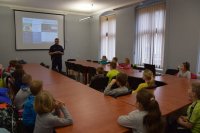 Zdjęcie przedstawia policjanta, który przypomina grupie dzieci podstawowe numery alarmowe.  Przedszkolaki siedzą w sali narad tarnogórskiej komendy