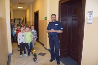 Zdjęcie przedstawia policjanta oraz grupę przedszkolaków idących korytarzem w tarnogórskiej komendzie