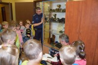 Zdjęcie przedstawia dwóch policjantów. Jeden jest umundurowany drugi, który klęczy nie jest umundurowany oraz grupę dzieci, które oglądają stare aparaty fotograficzne znajdujące się w  szklanej gablocie.