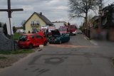 Zdjęcie przedstawia dwa pojazdy biorące udział w zdarzeniu drogowym. Samochody są rozbite. W tle widać wozy Straży Pożarnej oraz strażaków.