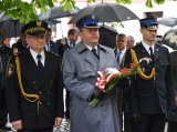 Zdjęcie przedstawia  Pierwszego Zastępcę Komendanta Powiatowego Policji w Tarnowskich Górach podinsp. Sławomira Bylickiego, który trzyma w dłoni wiązankę kwiatów. Obok niego z lewej strony znajduje się  Komendant Straży Miejskiej, a z prawej przedstawiciel Straży Pożarnej