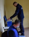 Zdjęcie przedstawia policjanta, który znajduje się przed urządzeniem służącym do wykonywania badania stanu trzeźwości. Na krześle siedzi mężczyzna - nietrzeźwy kierujący.