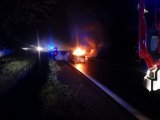 Zdjęcie przedstawia palący się samochód osobowy. W oddali widać policyjny radiowóz z włączonymi światłami błyskowymi oraz stojących przy nim policjantów.