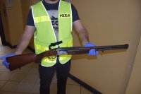zdjęcie przedstawia policjanta ubranego w kamizelkę odblaskową z napisem POLICJA, który trzyma w rękach broń myśliwską