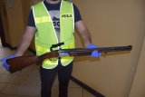 zdjęcie przedstawia policjanta ubranego w kamizelkę odblaskową z napisem POLICJA, który trzyma w rękach broń myśliwską
