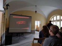 Zdjęcie przedstawia rzutnik, na którym widnieje napis # NAJGORZEJ. Z prawej strony fotografii widać grupę młodzieży oraz policjanta.