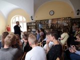 Zdjęcie przedstawia grupę młodzieży oraz policjanta. Policjant trzyma w dłoni mikrofon i prowadzi pogadankę.