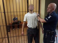 Z prawej strony fotografii widać policjanta, który trzyma za rękę mężczyznę - sprawcę kradzieży. Z lewej strony zdjęcia widać kraty. Za kratami siedzi mężczyzna - drugi ze sprawców.