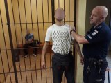 Z prawej strony fotografii widać policjanta, który trzyma za rękę mężczyznę - sprawcę kradzieży. Z lewej strony zdjęcia widać kraty. Za kratami siedzi mężczyzna - drugi ze sprawców.