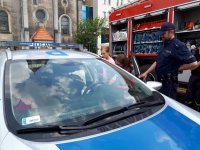 Zdjęcie przedstawia policyjny radiowóz, do którego wsiadają dzieci. Obok stoi policjant. W tle fotografii widać wóz strażacki.