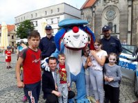 Zdjęcie przedstawia  maskotkę śląskiej policji psa &quot;Sznupka&quot;. Obok maskotki stoją dwaj umundurowani policjanci, grupa dzieci oraz organizator imprezy.