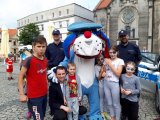 Zdjęcie przedstawia  maskotkę śląskiej policji psa &quot;Sznupka&quot;. Obok maskotki stoją dwaj umundurowani policjanci, grupa dzieci oraz organizator imprezy.