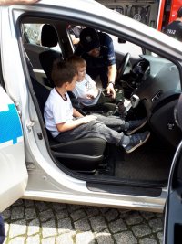 Zdjęcie przedstawia środek policyjnego radiowozu. Wewnątrz siedzi dwóch chłopców, widać również policjanta, który pokazuje dzieciom wyposażenie radiowozu.