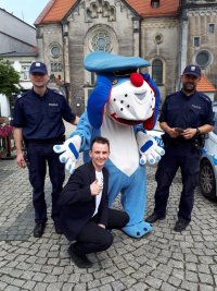 Zdjęcie przedstawia  maskotkę śląskiej policji psa &quot;Sznupka&quot;. Obok maskotki stoją dwaj umundurowani policjanci. Widać również kucającego mężczyznę - organizatora imprezy.
