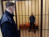 Z lewej strony fotografii widać policjanta obserwującego mężczyznę, który znajduje się za kratami.