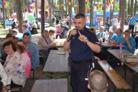 Na zdjęciu widać  policjanta asp. szt. Adama Cichowskiego oraz grupę siedzących przy stołach osób