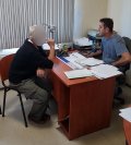Na zdjęciu widać dwóch mężczyzn siedzących po obu stronach biurka. Mężczyzna widoczny z prawej strony fotografii to policjant przesłuchujący mężczyznę widocznego z lewej strony fotografii.