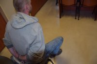 Na fotografii widać mężczyznę, który siedzi na krześle. Za plecami trzyma ręce, które są skute kajdankami.