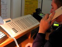 Fotografia kolorowa. Na zdjęciu widać siedzącego przy biurku policjanta, który trzyma przy uchu słuchawkę od aparatu telefonicznego.