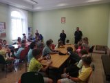 na zdjęciu widać grupę dzieci siedzących przy stołach. W tle widać policjantów
