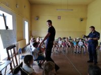 na zdjęciu widać grupę  siedzących na krzesłach dzieci. Pośrodku  nich stoją dwaj policjanci.
