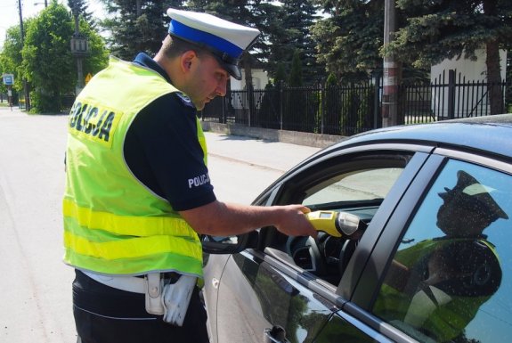zdjęcie przedstawia policjanta z wydziału ruchu drogowego, który w dłoni trzyma urządzenie do badania stanu trzeźwości i  bada nim kierowcę samochodu, który zatrzymał do kontroli