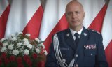 zdjęcie przedstawia Komendanta Głównego Policji generalnego inspektora Jarosława Szymczyka. Obok niego widać biało -czerwone kwiaty, a za nim biało-czerwone flagi