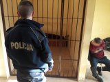 Na zdjęciu widać stojącego tyłem policjanta. Obok niego na krześle siedzi zakuty w kajdanki mężczyzna. W tle fotografii widać kraty, za którymi znajduje się drugi mężczyzna