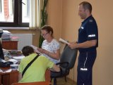 Z prawej strony fotografii widać umundurowanego policjanta. Z lewej strony zdjęcia widać siedzących przy biurku kobietę - policjantkę i odwróconego tyłem młodego mężczyznę.