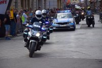 Na fotografii widać policjantów na motocyklach, policyjny radiowóz. Za nimi jedzie peleton kolarzy. Po obydwu stronach ulicy znajduje się tłum ludzi